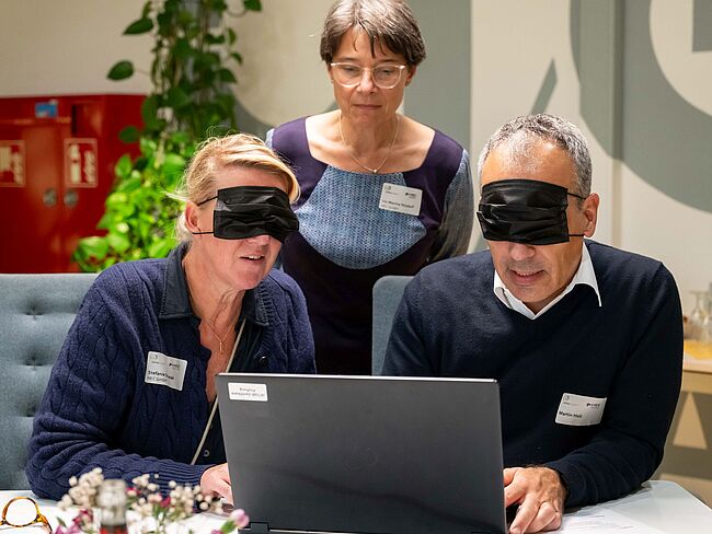 Eine Frau und ein Mann versuchen am Laptop, mit verbundenen Augen online ein Sofa zu bestellen.