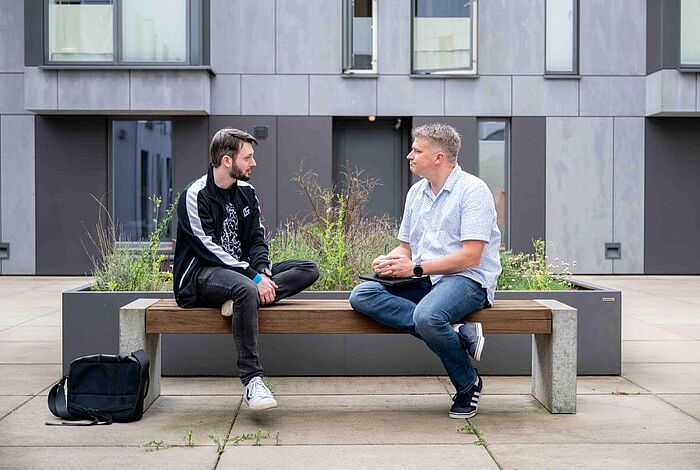 Kollegen sitzen auf einer Bank vor einem Beet und reden zugewandt