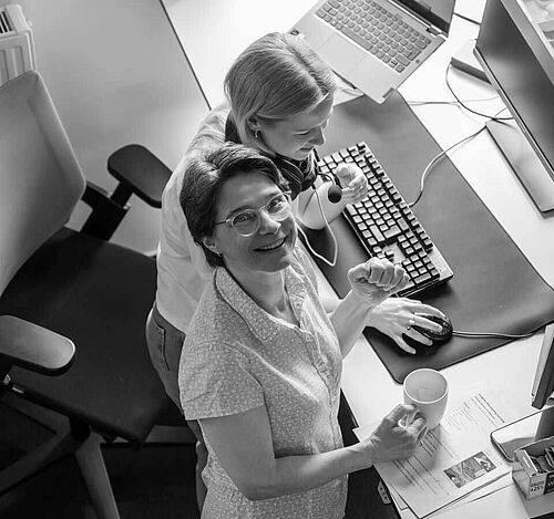 Zwei Frauen arbeiten gemeinsam stehend vor zwei Monitoren an einem Laptop am Schreibtisch im Büro