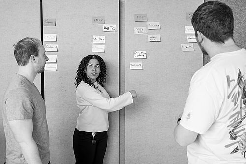 Drei Personen stehen im Büro der HEC und sehen auf einen Raumteiler, der mit beschriebenen Klebezetteln bestückt ist. 