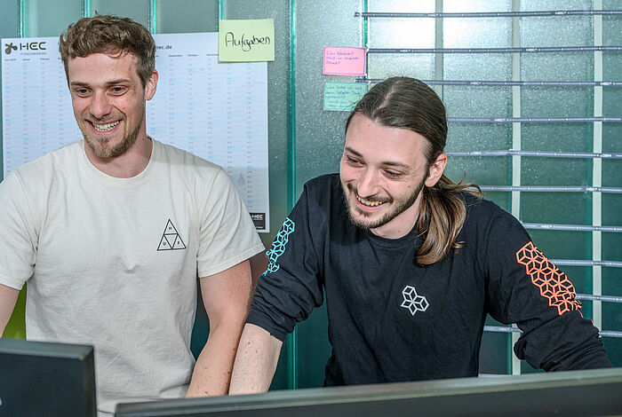 Zwei junge Männer stehend, lachen und blicken auf einen Monitor