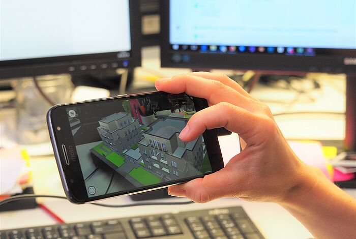 Eine Hand hält ein Smartphone, auf dem Virtuelle Gegenstände auf dem Schreibtisch zu sehen sind