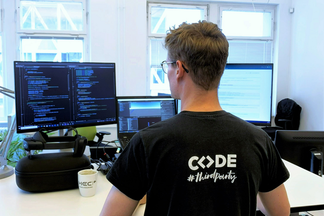 Werkstudent der HEC in Rückenansicht. Auf dem T-Shirt steht CODE. Er steht vor einem Schreibtisch. Werkstudent der HEC in Rückenansicht. Auf dem T-Shirt steht CODE. Er steht vor einem Schreibtisch.