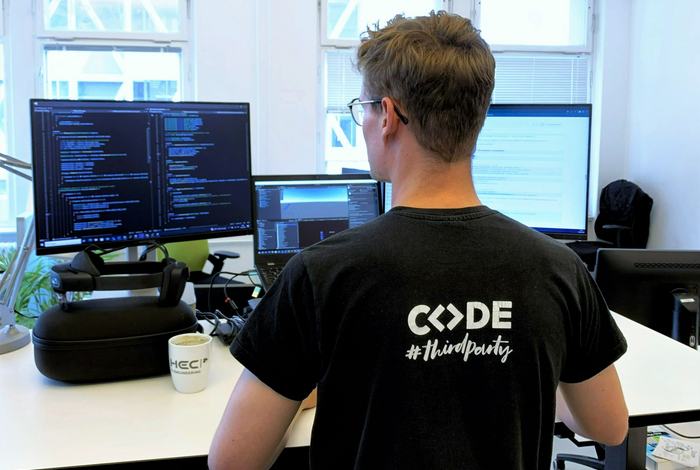 Paul Schilling, Werkstudent Werkstudent der HEC in Rückenansicht. Auf dem T-Shirt steht CODE. Er steht vor einem Schreibtisch.