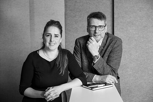 Eine Frau und ein Mann stützen sich auf einen Stehtisch und schauen in die Kamera.