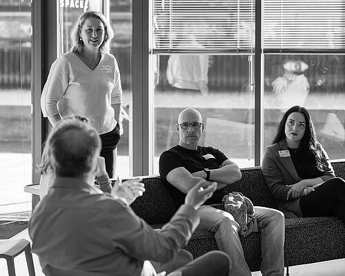 Zwei Frauen und zwei Männer sitzen auf einem Sofa oder stehen daneben.