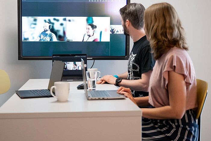 Ein Mann und eine Frau am Besprechungstisch sprechen digital mit anderen, die auf dem Großbildschirm zu sehen sind.