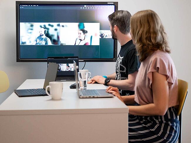 Ein Mann und eine Frau am Besprechungstisch sprechen digital mit anderen, die auf dem Großbildschirm zu sehen sind.