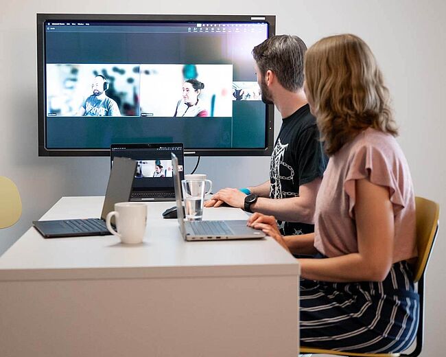Ein Mann und eine Frau am Besprechungstisch sprechen digital mit anderen, die auf dem Großbildschirm zu sehen sind.