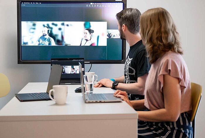 Ein Mann und eine Frau am Besprechungstisch sprechen digital mit anderen, die auf dem Großbildschirm zu sehen sind.