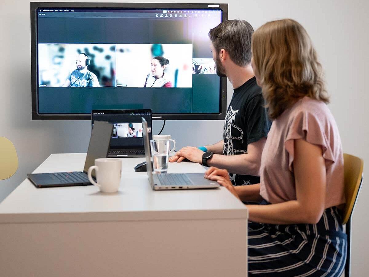 Ein Mann und eine Frau am Besprechungstisch sprechen digital mit anderen, die auf dem Großbildschirm zu sehen sind.