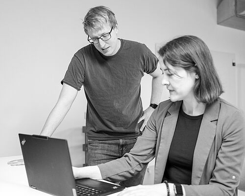 Eine Frau arbeitet am Laptop und zeit einem Mann, der neben ihr steht, etwas auf dem Bildschirm.