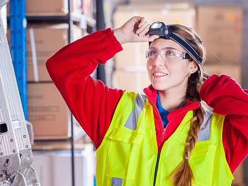 Auf dem Bild ist einer Arbeiterin in einer Firma zu erkennen. Sie trägt eine gelbe Warnweste und eine Schutzbrille. 